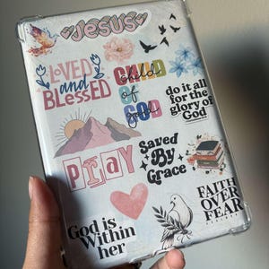 May include: A clear tablet case decorated with various stickers. The stickers include phrases like "Jesus," "Loved and Blessed," "Child of God," and "Saved By Grace." Other stickers feature a dove, flowers, and a heart. The case is held in a hand.