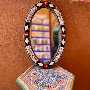 May include: An oval mirror with a black frame featuring white diamond shapes and red teardrop accents. The mirror sits atop a hexagonal pedestal decorated with intricate blue, pink, yellow, and green geometric floral patterns on its surface.
