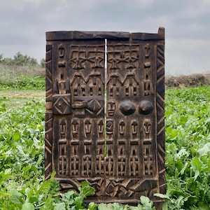 Könnte beinhalten: Eine dunkelbraune, aufwendig geschnitzte Holztür mit einem symmetrischen Design. Die Tür weist Reliefschnitzereien von menschlichen Figuren, geometrischen Formen und anderen symbolischen Elementen auf. Die Tür steht in einem Feld mit grünen Pflanzen unter einem bewölkten Himmel.