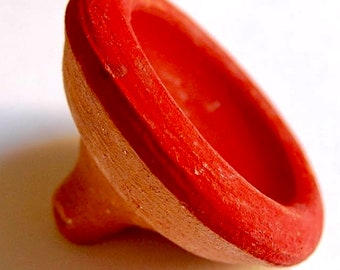 May include: A terracotta-colored, handmade clay object with a flared, bowl-like top and a small, rounded base. The rim is painted a vibrant red, contrasting with the natural clay color. The object is likely a small bowl or decorative piece.
