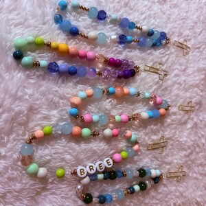 May include: Several beaded rosaries in various colors, including blue, green, pink, and purple. Each rosary features a gold-colored cross charm. One rosary has the name "BREE" spelled out in white letter beads.