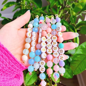 May include: A set of colorful beaded bracelets with gold lettering spelling out the words "BABY", "PUSH", "RN", and "OB".