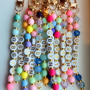 May include: A collection of beaded keychains featuring gold-tone lobster clasps and colourful acrylic beads. Each keychain includes white round beads with gold lettering spelling out OBGYN, interspersed with solid, translucent, and glittery beads.