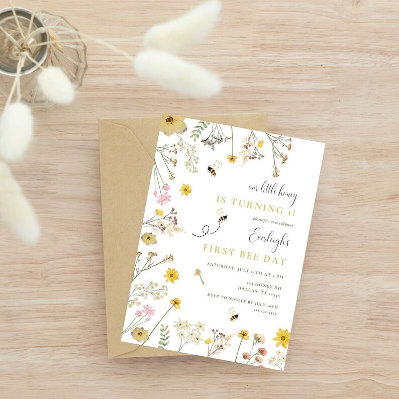 Puede incluir: Una tarjeta de invitaci&oacute;n blanca con un borde floral y un dise&ntilde;o de abeja. El texto en la tarjeta dice "&iexcl;nuestro peque&ntilde;o miel cumple 1!" y "&iexcl;Primer d&iacute;a de la abeja!".