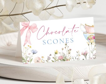 Puede incluir: Una tarjeta blanca plegada con las palabras "Chocolate Scones" en escritura rosa y azul. Un lazo rosa e ilustraciones florales decoran el lado izquierdo. Tazas de té y más flores están a la derecha. La tarjeta está sobre una bandeja de madera blanca.