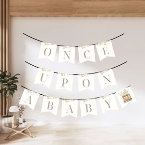 Puede incluir: Una pancarta blanca con las palabras "Once Upon A Baby" en letras grises. La pancarta está decorada con pequeños diseños florales y está colgada de una cuerda negra.