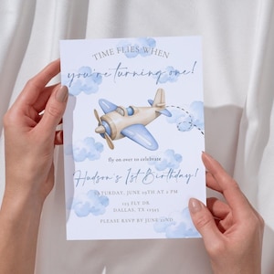 May include: A light blue and beige airplane illustration on a white invitation card. The card reads "Time flies when you're turning one!" and "Hudson's 1st Birthday!" with additional event details. The invitation is held by two hands.