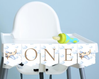 Könnte beinhalten: Ein weißer Hochstuhl mit einem Banner mit der Aufschrift "ONE" in braunen Buchstaben. Das Banner zeigt Flugzeug- und Wolkenillustrationen. Ein blau-grüner Babynahrungszubereiter liegt auf dem Tablett. Der Hintergrund ist hellblau.