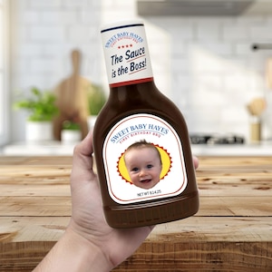 以下が含まれることがあります： 赤ちゃんの写真と「Sweet Baby Hayes First Birthday BBQ」の文字が入ったカスタムラベルが付いた、茶色のバーベキューソースのボトル。「The Sauce is the Boss!」と「NET WT 230g」の文字も入っています。