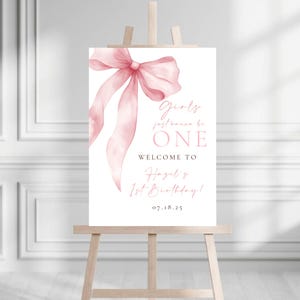 May include: A white welcome sign with a pink watercolor bow and the text "Girls just wanna be ONE. Welcome to Hazel's 1st Birthday! 07.18.25"