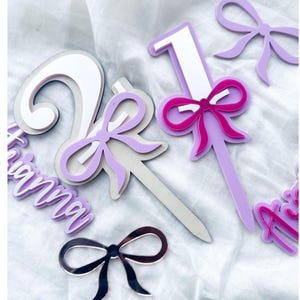 May include: Purple and white acrylic cake toppers featuring the numbers 1 and 2, a pink bow, and the name "Gianna" in purple. The toppers are displayed against a white background.