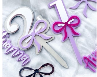 May include: Purple and white acrylic cake toppers with the numbers 1 and 2, a pink bow, and the name "Gianna" in purple. The toppers are on a white background.