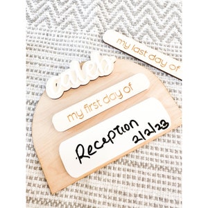 Op de afbeelding: Houten bord met de naam "Caleb" in witte letters. Het bord heeft twee witte rechthoekige labels met gouden tekst die "my first day of" en "my last day of" zeggen. Er is ook een wit rechthoekig label met zwarte tekst dat "Reception 21223" zegt.
