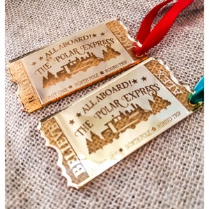 May include: Two gold-coloured ticket ornaments with the text "All Aboard! The Polar Express" and "Believe" on them. The tickets are decorated with a train and trees.