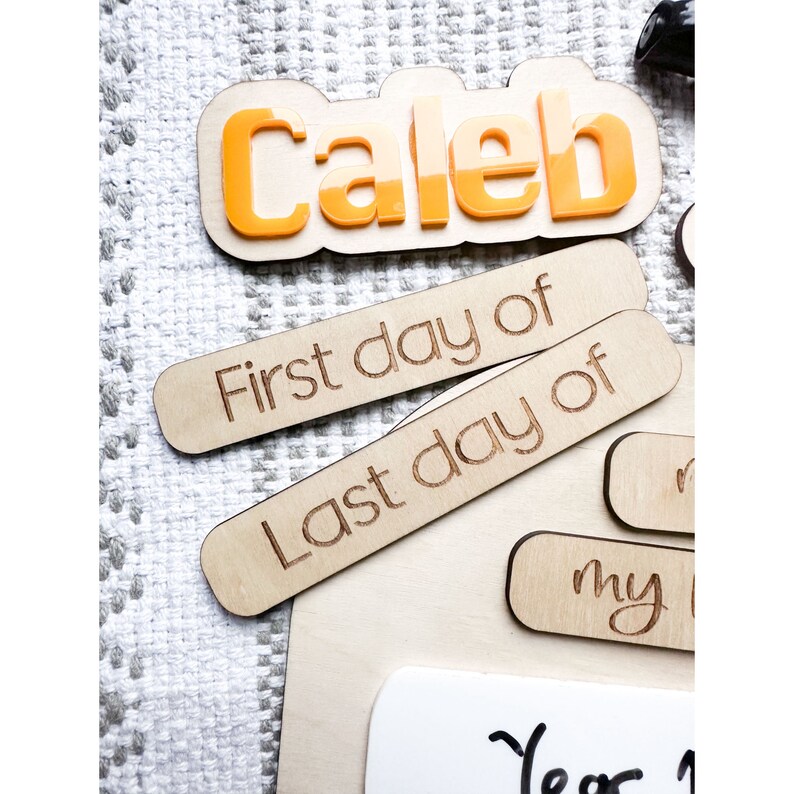 Op de afbeelding: Houten bord met de naam "Caleb" in oranje letters. Het bord heeft ook twee houten stukken die "Eerste dag van" en "Laatste dag van" zeggen.
