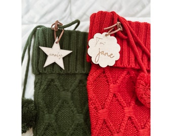 May include: Two knitted Christmas stockings with personalized wooden tags. The stocking on the left is green with a star-shaped tag that says "carly". The stocking on the right is red with a round tag that says "To: jane".