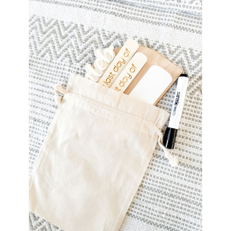Op de afbeelding: Een witte canvas tas met een houten bord waarop 'laatste dag van' staat en een houten bord waarop 'de laatste dag van' staat. Er is ook een wit droog uitwisbaar bord en een zwarte droog uitwisbare marker.