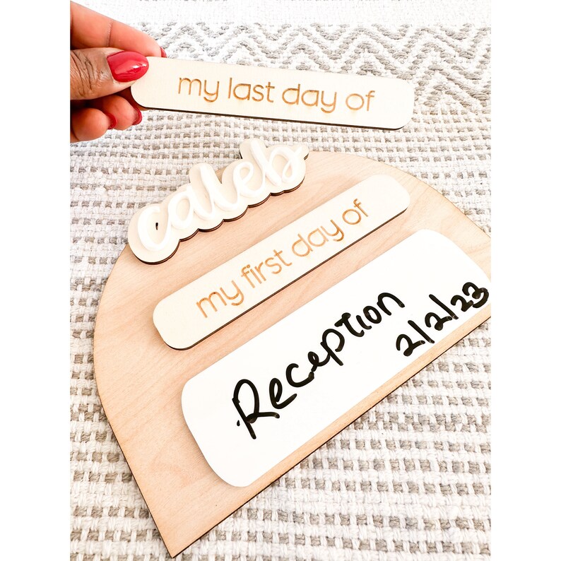 Op de afbeelding: Houten bord met een witte cursieve naam "Caleb" en drie witte rechthoekige borden. Het bovenste bord zegt "my last day of", het middelste bord zegt "my first day of" en het onderste bord zegt "Reception 2/21/23".