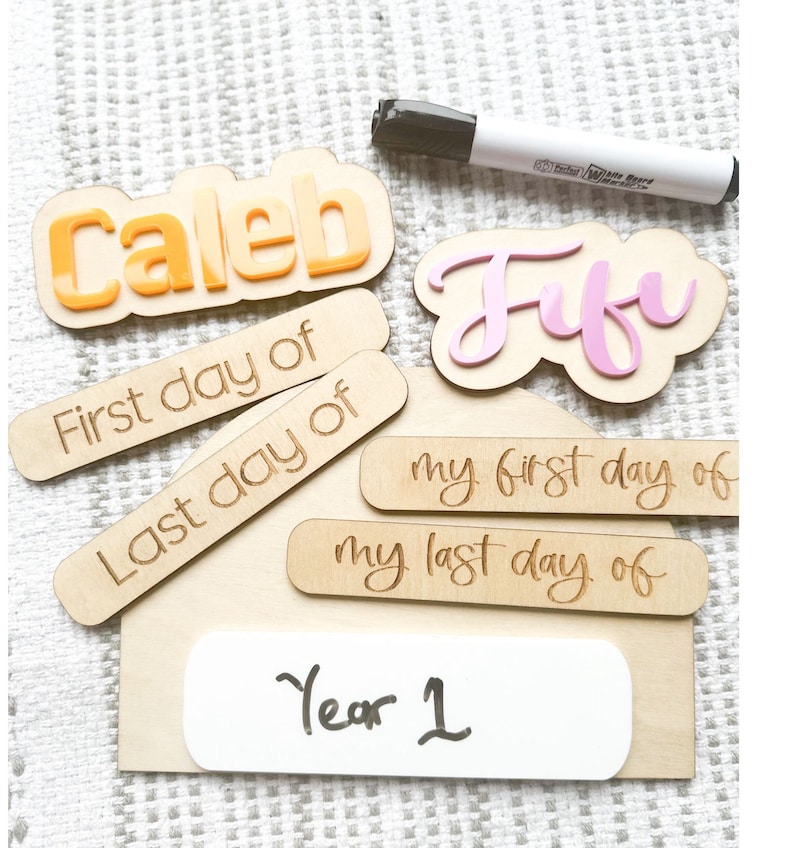 Op de afbeelding: Houten bord met de namen "Caleb" en "Jufu" in roze en oranje letters. Het bord bevat ook de zinnen "Eerste dag van", "Laatste dag van", "Mijn eerste dag van", "Mijn laatste dag van" en "Jaar 1".