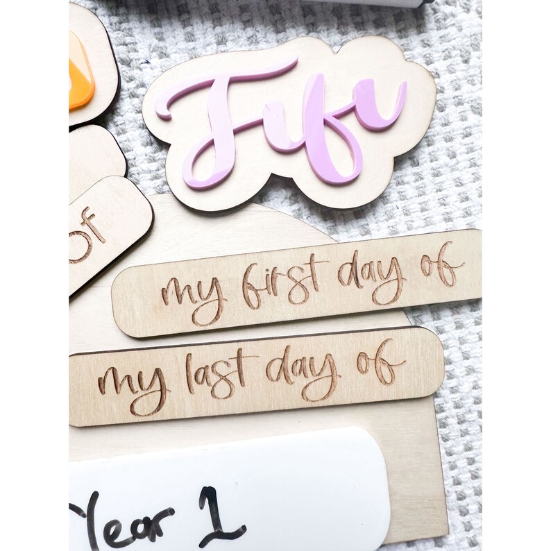 Op de afbeelding: Houten borden met de tekst "my first day of" en "my last day of" in een cursief schrift. Een roze houten bord met de naam "Fufu" in een cursief schrift. Een wit bord met de tekst "Year 1" erop.
