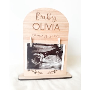 May include: Wooden baby announcement sign with the name "OLIVIA" and the phrase "coming soon" engraved. It displays a sonogram image, held by two small pegs. A floral design decorates the bottom of the sonogram.