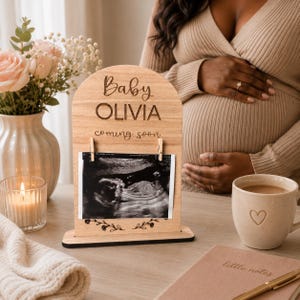 以下が含まれることがあります： Baby OLIVIA coming soonと刻印されたアーチ型の木製サイン。サインには2つの小さな木製クリップが付いており、白黒の超音波写真を保持しています。ベースは長方形で、木材は明るいナチュラルな仕上げになっています。