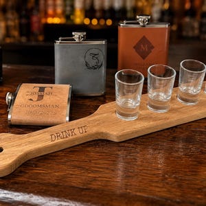 Pode incluir: Uma tábua de servir em madeira com o texto DRINK UP gravado na superfície, contendo quatro copos de shot de vidro transparente. Três cantis de metal estão posicionados atrás, incluindo um cinzento, um castanho e um bege com texto.