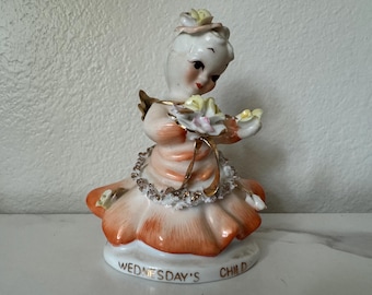 May include: A porcelain figurine of a girl in a peach-colored dress with a gold trim and a flower crown. She is holding a bouquet of flowers and the base of the figurine reads "Wednesday's Child".