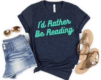 May include: A navy blue t-shirt with the text "I'd Rather Be Reading" in teal green lettering. The shirt is laid flat on a white background with a pair of denim shorts, sunglasses, and sandals.