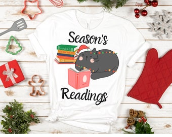 Op de afbeelding: Wit T-shirt met de tekst "Season's Readings" en een afbeelding van een grijze kat met een kerstmuts en lichtjes, die een boek leest. Feestelijke decoraties omringen het shirt.