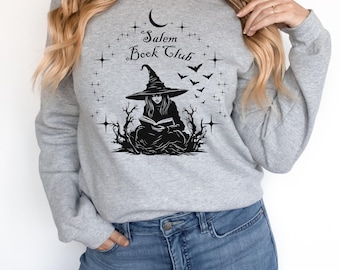 Könnte beinhalten: Hellgraues Sweatshirt mit dem Text "Salem Book Club" und einer schwarzen Grafik einer Hexe, die ein Buch liest. Die Hexe trägt einen spitzen Hut und ist von Fledermäusen, Sternen und einem Halbmond umgeben.