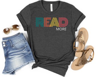 Puede incluir: Una camiseta gris oscuro jaspeada con un gráfico retro de colores "LEE MÁS".
