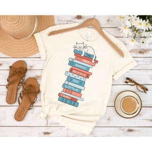 May include: A white t-shirt with a graphic of a cat sleeping on a stack of books. The books have the words "So Many Books So Little Time" written on them.
