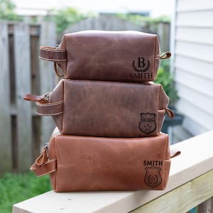 May include: Three stacked brown leather toiletry bags. The top bag has the letter "B" inside a wreath and the name "SMITH". The middle bag has a badge graphic. The bottom bag has the name "SMITH" and a badge graphic. Each bag has a handle.