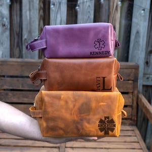 May include: Three leather toiletry bags in purple, brown, and orange are stacked. The purple bag has a medical symbol and the name "Kennedy". The brown bag has the name "Liam". The orange bag has a medical symbol and the name "Anderson".