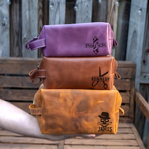 May include: Three leather toiletry bags in purple, brown, and orange. Each bag has a handle and a black graphic with a name. The top bag says "Patrick" with a barber pole. The middle bag says "Kennedy" with scissors. The bottom bag says "James" with a skull.