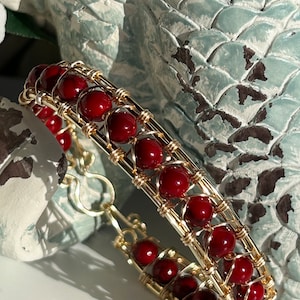 May include: A gold-toned wire-wrapped bracelet featuring rows of deep red beads. The beads are evenly spaced and create a textured, eye-catching design. The bracelet has a secure clasp.