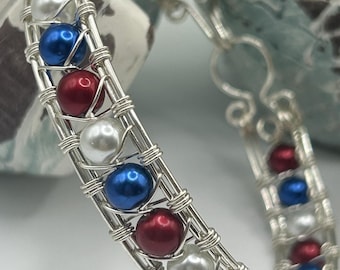 Puede incluir: Un brazalete de alambre plateado envuelto con cuentas rojas, blancas y azules. El brazalete está hecho con un diseño de alambre tejido.