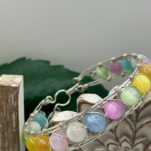 May include: A handmade bracelet featuring colorful, round beads in shades of yellow, orange, pink, blue, green, and white. The beads are strung on a silver wire, creating a unique and eye-catching accessory. The bracelet is designed with a clasp for easy wear.