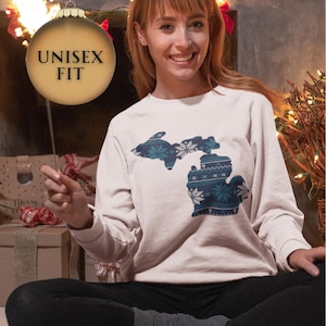 May include: A light pink unisex sweatshirt featuring a detailed blue and white design of the state of Michigan. The design incorporates snowflake and geometric patterns. The sweatshirt has long sleeves and a crew neck. The text "UNISEX FIT" is on a gold ornament.