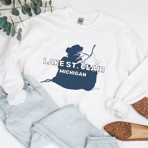 May include: White sweatshirt with a navy blue graphic of Lake St. Clair and the text "LAKE ST. CLAIR MICHIGAN". The sweatshirt is paired with light blue jeans and leopard print flats.
