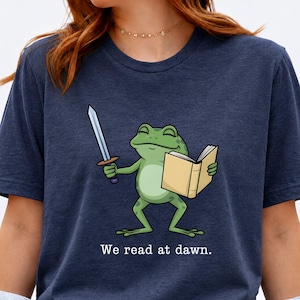 May include: A dark blue t-shirt featuring a graphic of a green frog holding a silver sword in one hand and an open book in the other. Below the frog, the text reads, "We read at dawn." The shirt has a crew neckline and short sleeves.