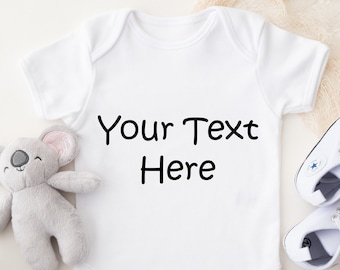 May include: A white baby bodysuit with the text "Your Text Here" in black. The image includes a grey koala soft toy, a pair of white baby trainers, and some greenery. The bodysuit is a short-sleeved garment.