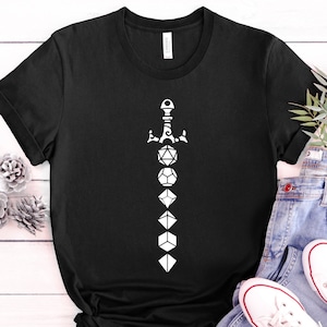 Peut inclure: T-shirt noir avec un graphique blanc d'une épée faite de dés. L'épée est composée de sept dés, chacun avec un nombre différent de points.