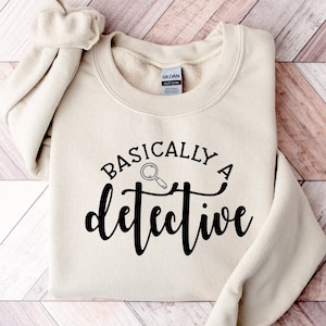 May include: A beige sweatshirt with the text "BASICALLY A detective" in black lettering with a magnifying glass icon.