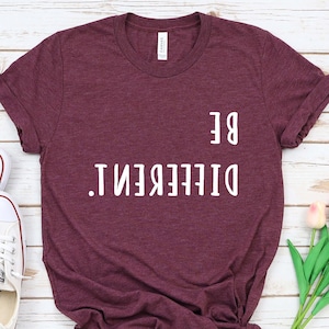 Peut inclure: Un t-shirt marron avec le texte "BE DIFFERENT." imprimé en blanc sur le devant. Le t-shirt est posé à plat sur une surface en bois blanc avec une paire de baskets blanches et un bouquet de tulipes roses.