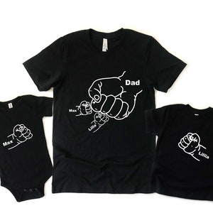 Peut inclure: T-shirts noirs avec des contours blancs de mains se tenant l'une l'autre. Le t-shirt adulte dit "Papa" et les t-shirts enfants disent "Max" et "Lillia".