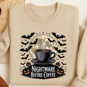 May include: Beige sweatshirt with a graphic featuring a coffee cup, bats, pumpkins, and a full moon. The text "Nightmare Before Coffee" is below the cup. The design is in black and white.