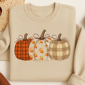 Peut inclure: Sweat-shirt beige avec trois citrouilles décoratives. Les citrouilles présentent différents motifs : carreaux orange, floral orange sur fond blanc et carreaux beige. Les citrouilles sont disposées en rangée, parfaites pour l'automne.