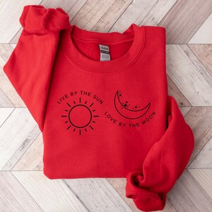 May include: Red sweatshirt with a black graphic design that says "Live by the sun, love by the moon" with a sun and moon illustration.